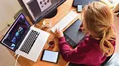 A person with long hair is sitting at a desk and using a graphic tablet. Around them, theres a laptop, a smartphone, a monitor, and a glass of water. The area looks creative and full of tech gadgets.
