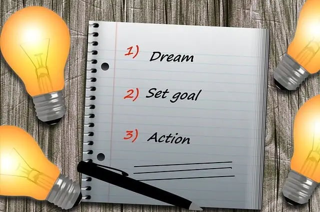 A notepad with a list that says: 1) Dream, 2) Set a goal, 3) Take action next to a pen. Its on a wooden background, and there are light bulbs around the notepad to show ideas and inspiration.