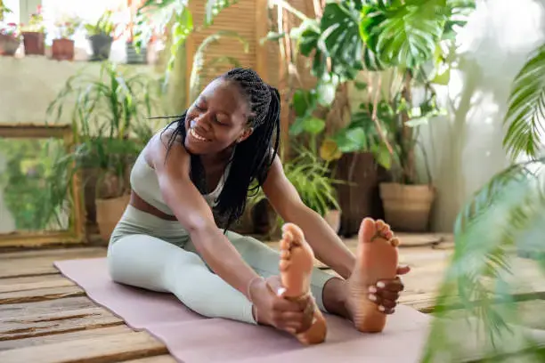 A person with long hair is doing yoga and smiling while sitting on a pink mat in a sunny room with lots of green plants around. Enjoying fitness and staying healthy, they are wearing a light green workout outfit and reaching forward to touch their toes.
