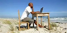 Someone is sitting at a wooden desk on a sandy beach, using a laptop. The beach has patches of dune grass here and there. In the background, you can see ocean waves under a clear blue sky.