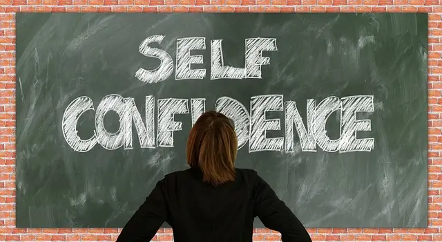 A person with brown hair is standing in front of a chalkboard that has Self Confidence written on it in big white letters. This shows they have an entrepreneurs mindset. The background is a brick wall, making it feel like theyre planning out big goals. We cant see the persons face because their back is turned to us.