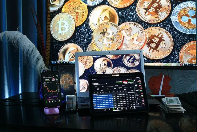 A cool desk setup includes a computer, tablet, and smartphone showing cryptocurrency charts. In the background, there’s a big screen with Bitcoin and other digital currencies. A small pile of cash suggests investment ideas, and theres a plant to add some greenery to this trading space.