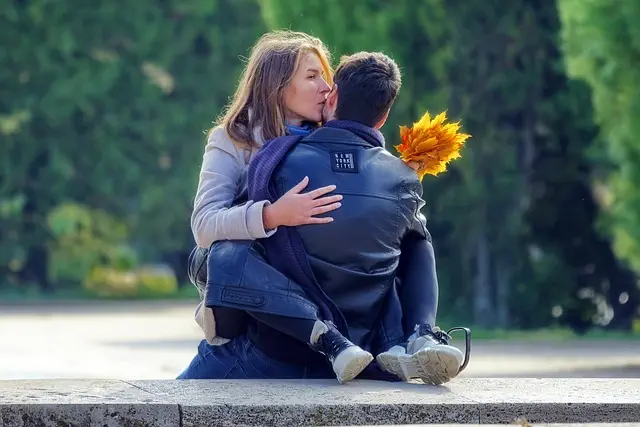 A couple is sitting together outside on a ledge, hugging peacefully. The woman has a bunch of yellow leaves in her hands and is leaning in to kiss the mans cheek. They are dressed casually, which adds to the calm and relaxed feeling of the moment. Behind them, there are green plants that make everything seem even more peaceful.