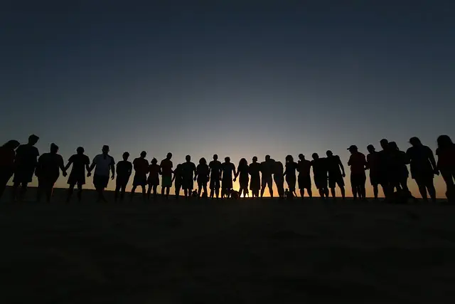 Against the backdrop of a sunset, a group of people hold hands, showing the spirit of being part of a helpful community. As the sun goes down, the sky changes from dark blue to orange and their shadows stretch across the sand. This reminds us how important it is to have supportive relationships in life.